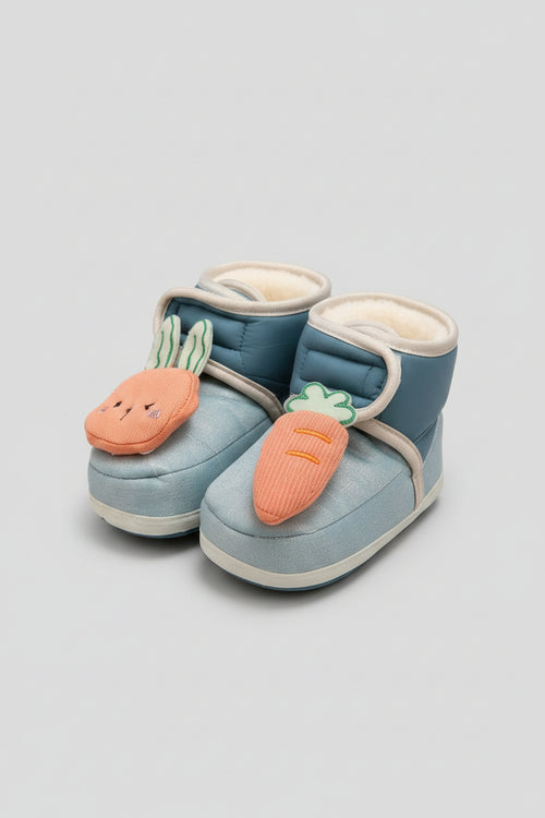 A pair of baby booties in light blue with plush cream lining, decorated with embroidered orange carrot and bunny face appliqués, shown on a soft beige textured surface with baby props.